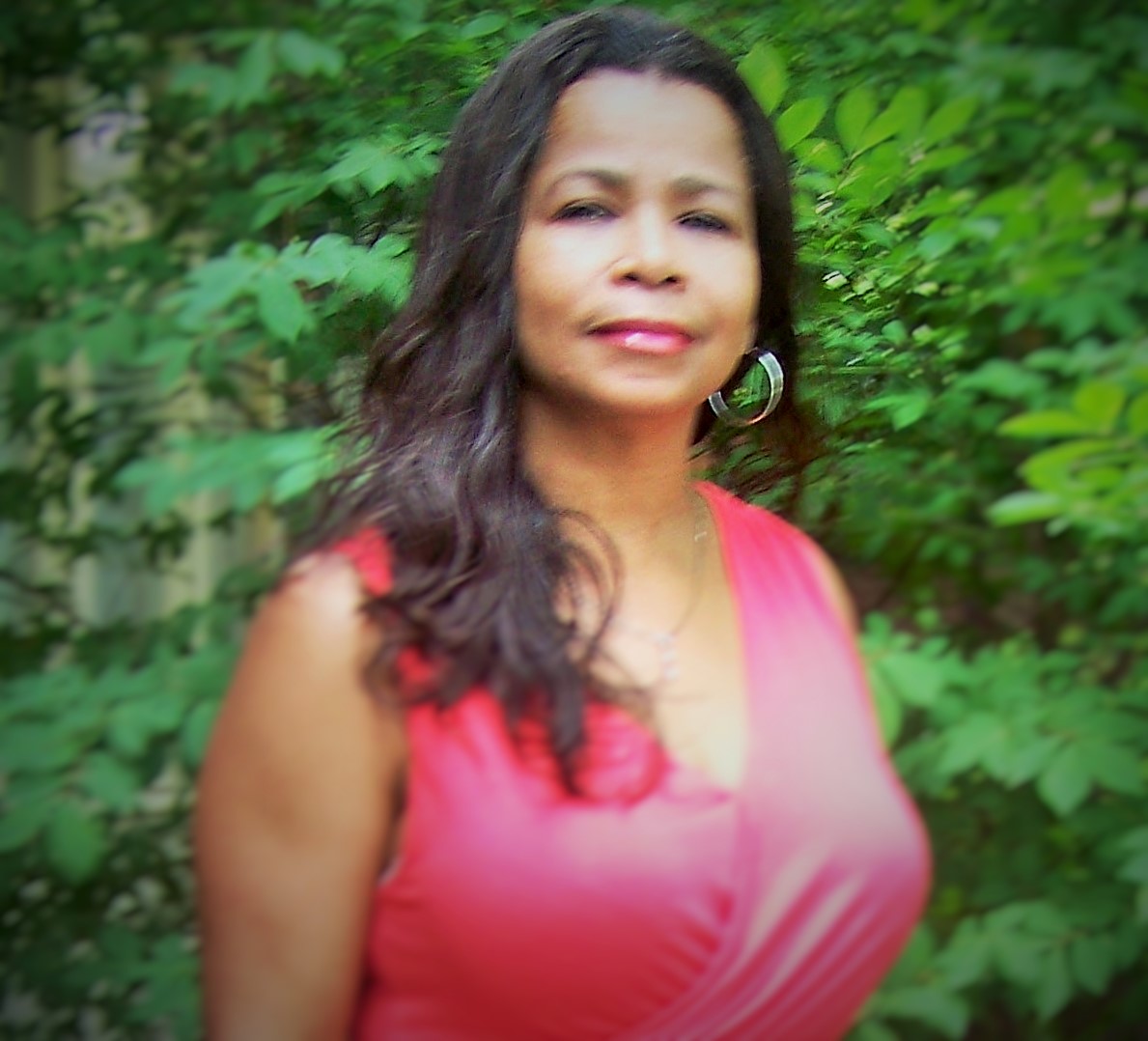 A woman with long, dark hair wearing a pink dress stands in front of lush green foliage.