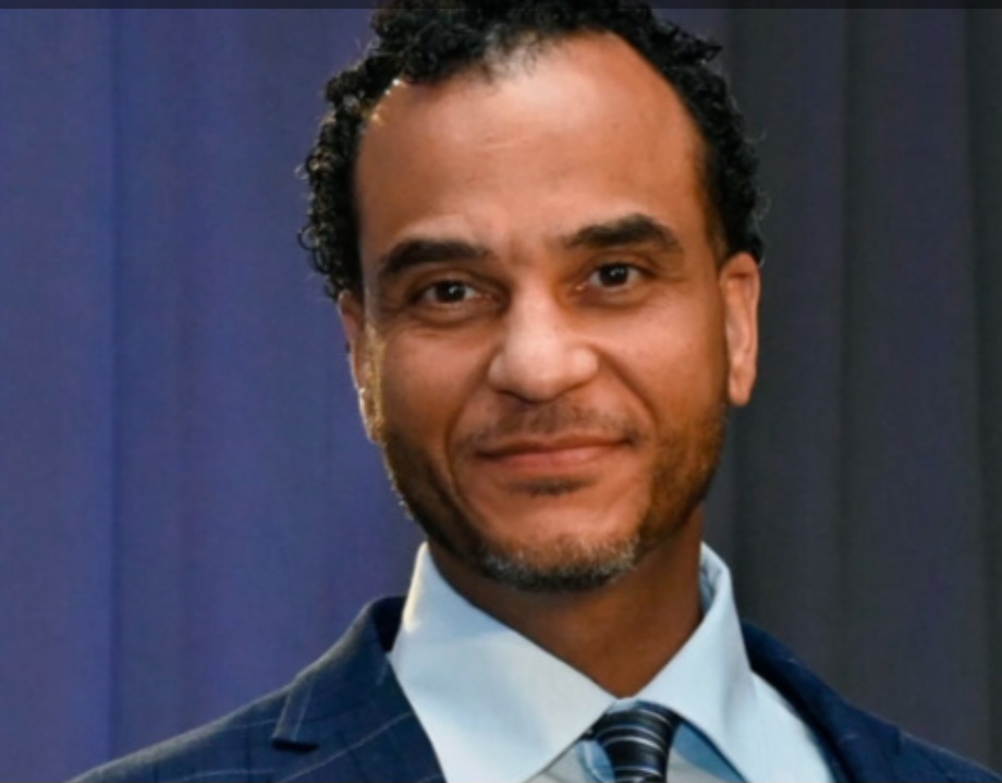 A man smiling in a suit with a tie, standing against a neutral-colored background.